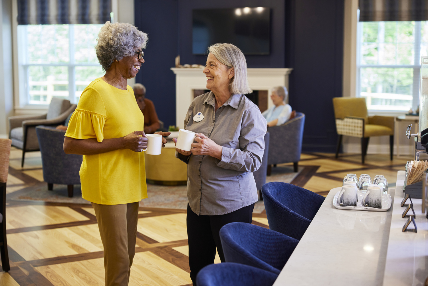 Staff member and resident holding mugs of coffee by a bar area with blue chairs